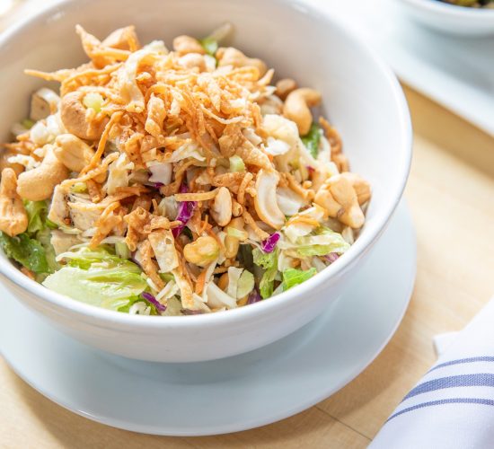 A bowl of salad topped with nuts, chopped vegetables, and crispy fried onions, placed on a wooden table. A folded cloth napkin is beside it, and another dish is partially visible in the background.