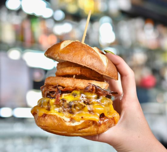 A hand holding a large, stacked cheeseburger with a pretzel bun. The burger is filled with melted cheese, bacon, and onion rings, skewered with a toothpick. The background features a blurred display of bottles and glasses.