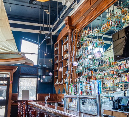 An upscale bar interior with a long wooden counter, high shelves filled with various liquor bottles, a large mirror behind the bar, and a ladder for access. The ceiling has an elegant design with hanging lights and a canvas fixture.