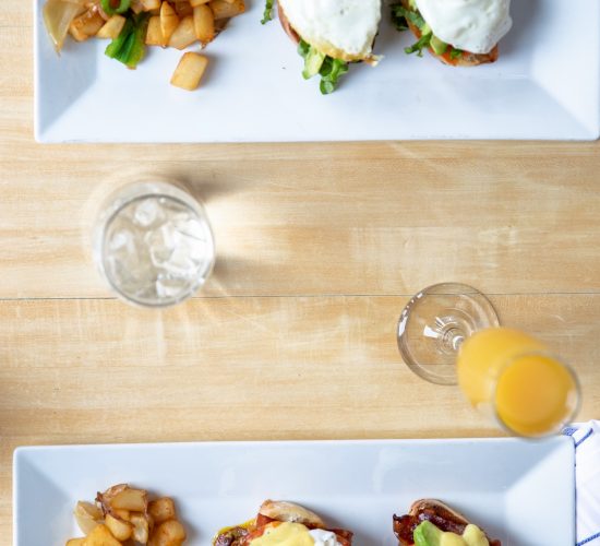 Overhead view of a wooden table with two plates. One plate has sunny-side-up eggs on bread with potato cubes. The other plate has eggs benedict with hollandaise sauce and potatoes. A glass of water and an orange drink are beside the plates.