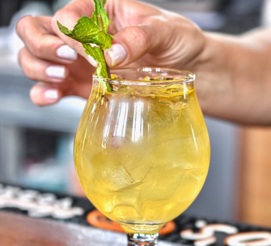 A hand garnishes a yellow iced cocktail with fresh mint leaves in a goblet-shaped glass, set on a wooden bar top with a blurred background.