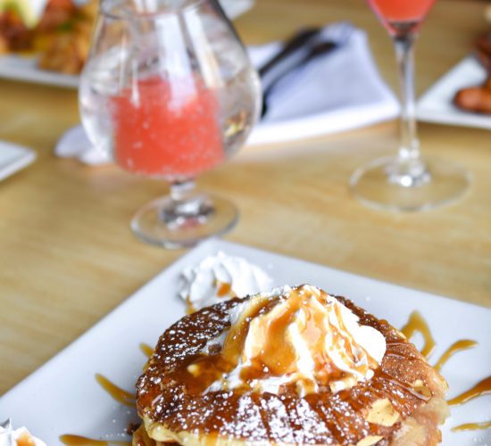 A stack of pancakes topped with whipped cream and caramel sauce on a white plate, garnished with whipped cream on the sides. In the background, a glass of pink cocktail and another with a similar drink rest on a wooden table.