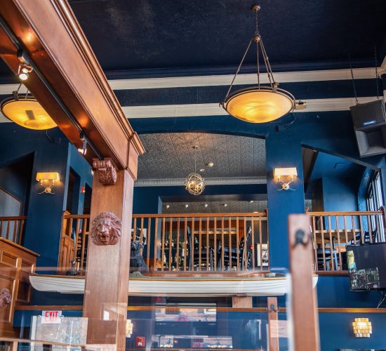 A modern restaurant interior featuring blue walls, a high ceiling, and warm lighting. Wooden railings enclose an upper mezzanine level with seating. A large TV screen is visible on the right side, and decorative chandeliers hang from the ceiling.