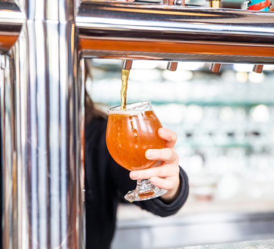 A person is pouring beer from a tap into a rounded glass at a bar. Several beer taps are visible, each with different handles. The background features blurred shelves with glassware.