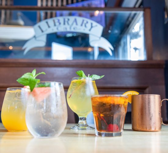 A variety of cocktails served on a wooden table in a bar setting. The background features a glass partition with the word "Library" visible. The drinks include a copper mug and glasses garnished with mint and fruit.