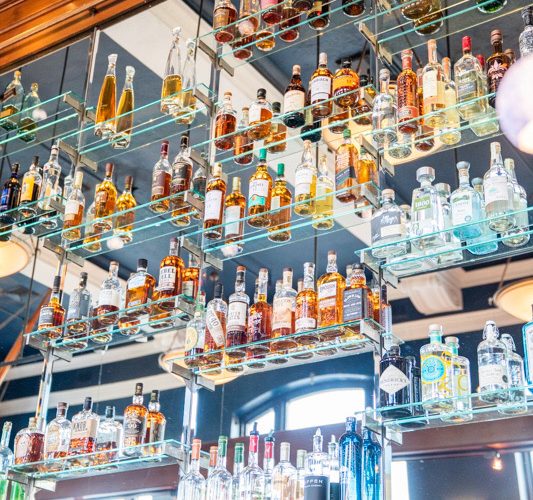 A wall with multiple glass shelves displaying a variety of liquor bottles, including whiskey, vodka, and gin. The setup is in a well-lit bar, with reflections from the ceiling and decor complementing the vibrant colors of the bottles.