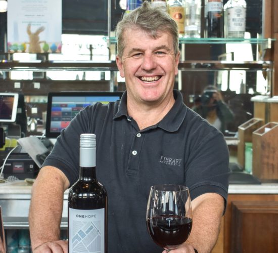 A smiling man stands behind a bar, holding a glass of red wine in one hand and a bottle in the other. Behind him, there are various liquor bottles on shelves. He is wearing a dark polo shirt.