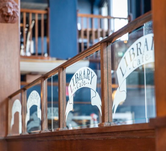 Interior of a cafe featuring wooden dividers with frosted glass panels that have "Library Cafe" written on them. The atmosphere is cozy, with soft lighting and visible bookshelves in the background.