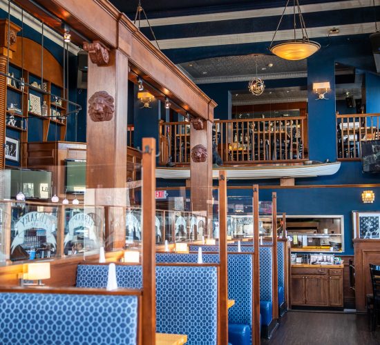 A stylish restaurant interior featuring blue and wood-themed decor. Booths with patterned blue upholstery line the right side, while a second-floor balcony overlooks the spacious area. Warm lighting and wooden accents create a cozy atmosphere.