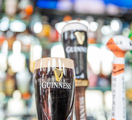 A pint of Guinness sits on a bar counter with foam at the top. Several beer taps are visible in the background, reflecting a pub setting. A blurry TV screen is mounted above the scene.