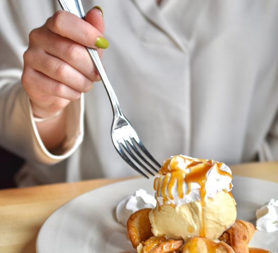 A person in a white shirt holds a fork above a dessert of ice cream topped with caramel sauce on a pastry, surrounded by whipped cream dollops on a white plate.