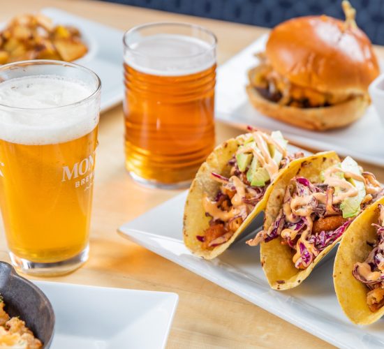 A table with a variety of foods, featuring three tacos topped with avocado and sauce, two burgers, sweet potato fries, a dish of pasta, and two glasses of beer. The setting is bright and inviting.