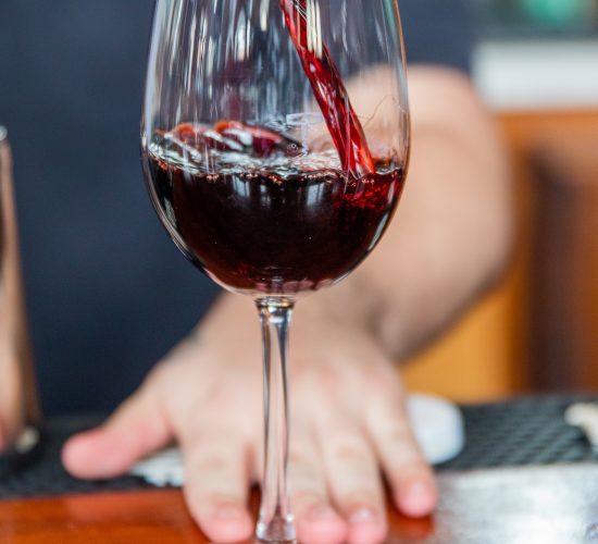 A close-up of a wine glass being filled with red wine. The glass is on a wooden surface, and a hand is visible in the background. The wine is mid-pour, creating a dynamic effect.