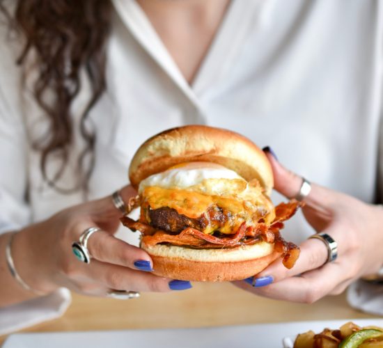 A person with curly hair holds a burger with bacon, cheese, and a poached egg. They have painted nails and are wearing rings. A side of fries is partially visible on the table. The person is wearing a white shirt.