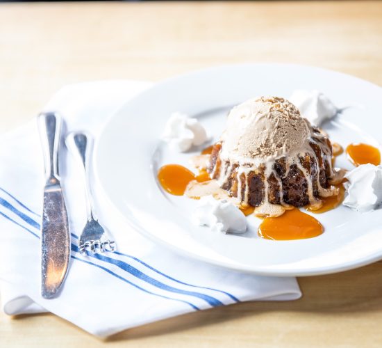 A dessert plate featuring a scoop of ice cream atop a rich cake drizzled with caramel sauce. Dollops of whipped cream surround the cake. The plate is set on a wooden table next to a blue-striped napkin with a knife and fork.