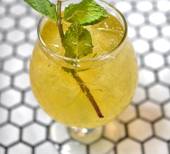 A refreshing iced drink with a light yellow hue, served in a round glass on a hexagonal white and black tile surface. The drink is garnished with a sprig of fresh mint, adding a touch of green.