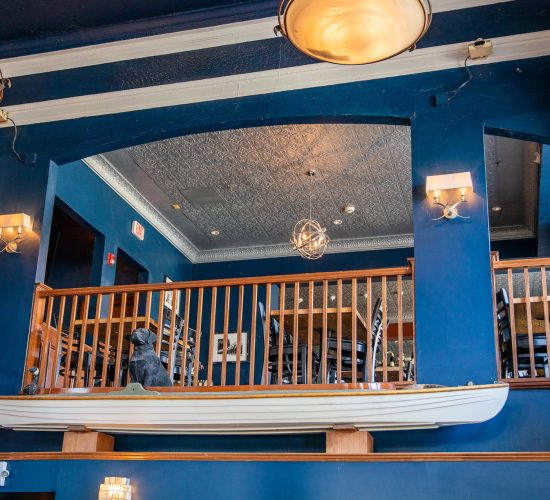 A cozy restaurant interior with navy blue walls, featuring a wooden railing overlooking a lower level. A decorative canoe is mounted near the ceiling, and a sculpted dog is placed at the railing. Warm lighting from sconces and a chandelier illuminates the space.