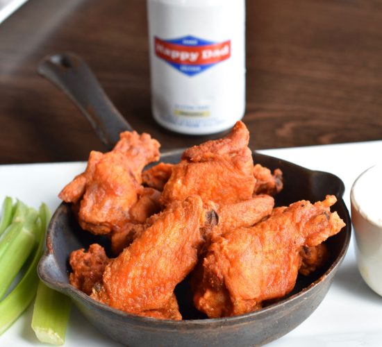 A skillet filled with crispy buffalo wings sits on a rectangular white plate. Celery sticks and a small bowl of dipping sauce are beside it. In the background, there is an unopened can labeled "Happy Dad." The table is a dark wood.