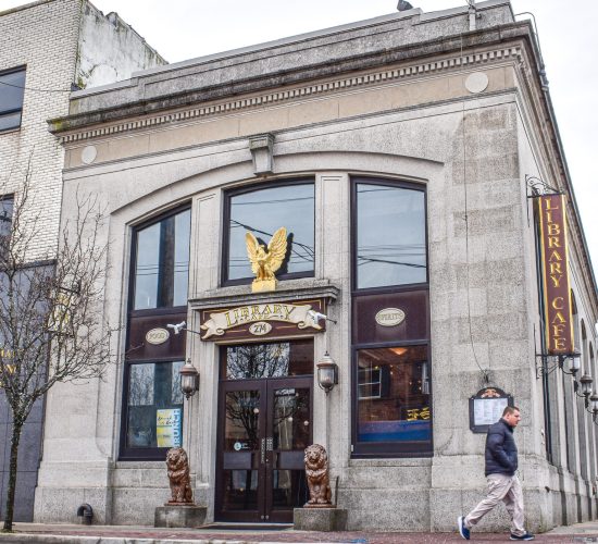 A small, historic stone building with large arched windows and a golden eagle ornament above the entrance. There are two lion statues at the doorway. A person walks by on the sidewalk. Sign reads "Library Cafe.