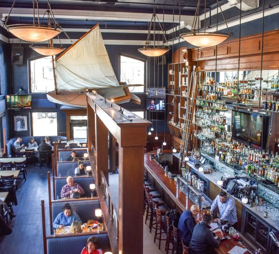 A lively restaurant with a maritime theme features a large sailboat model hanging from the ceiling. Patrons sit in booths and at the bar, surrounded by shelves stocked with various bottles. Natural light pours in through tall windows.