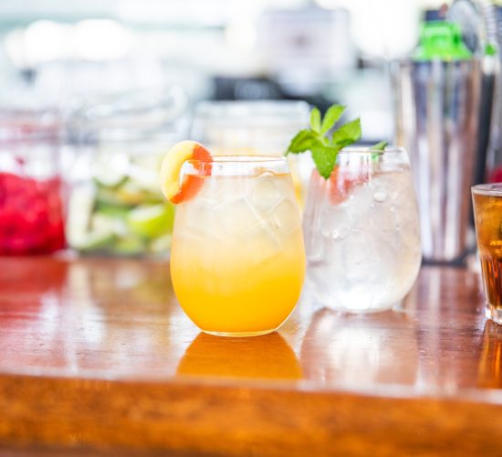 A refreshing cocktail garnished with a lemon slice on a wooden bar counter. Behind it are jars of fruits, a glass with mint leaves, and bottles. The background is softly blurred, emphasizing the drink in focus.