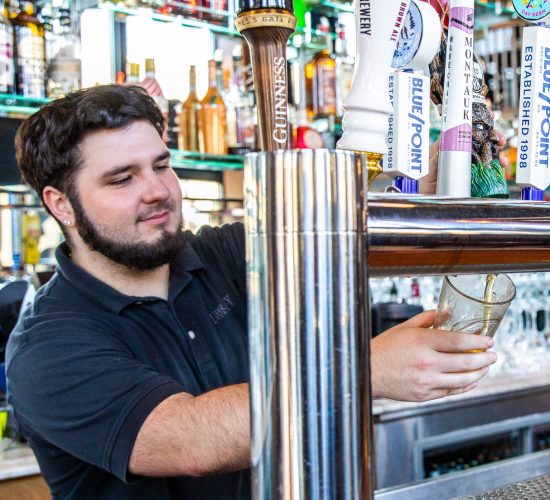 A bartender with a beard and wearing a black shirt is pouring a beer from a tap behind a bar. Various beer taps are visible, including brands like Guinness and Blue Point. The bar is stocked with assorted bottles in the background.