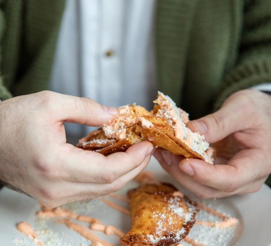 A person in a green sweater is breaking apart a fried pastry on a white plate. The plate has two pastries with powdered sugar and drizzle on top. The person is wearing a white shirt underneath the sweater.