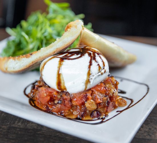 A gourmet dish featuring a poached egg on a bed of diced tomatoes and raisins, drizzled with balsamic glaze, is served on a white plate with lightly toasted bread and a side of fresh greens.