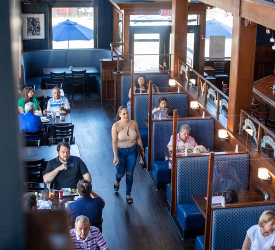 A restaurant interior with blue booth seating and wooden accents. Diners are enjoying meals, and a staff member walks through the aisle. Sailboat decor hangs above, and natural light comes from a large window in the background.