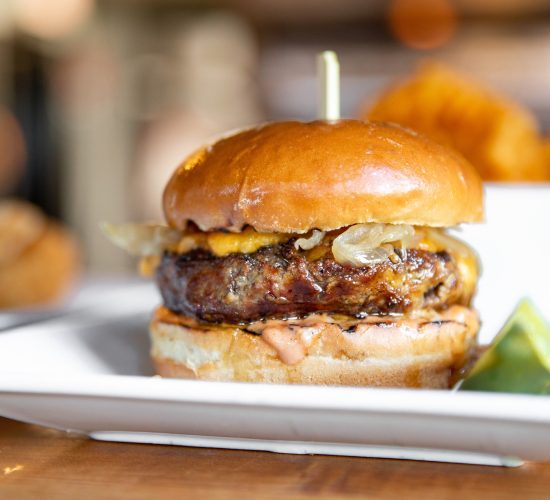 Close-up of a juicy burger with a glossy bun, melted cheese, and onions on a white plate. A lime wedge is on the side, and crinkle-cut fries are blurred in the background, creating a delicious and inviting scene.