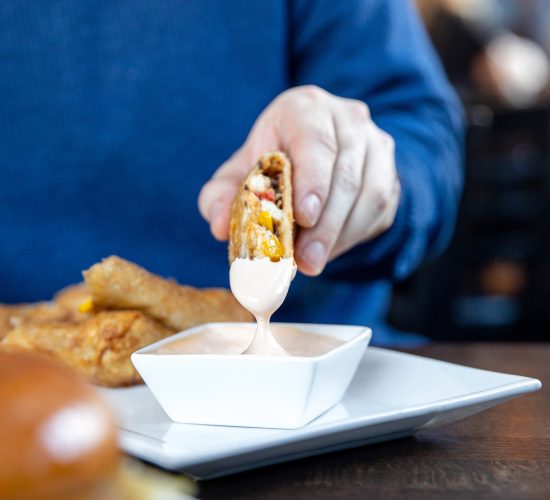 A person in a blue sweater dips a rolled taco into a square bowl of creamy dipping sauce. In the foreground, a burger and fries are visible on a plate. The restaurant setting is slightly blurred in the background.