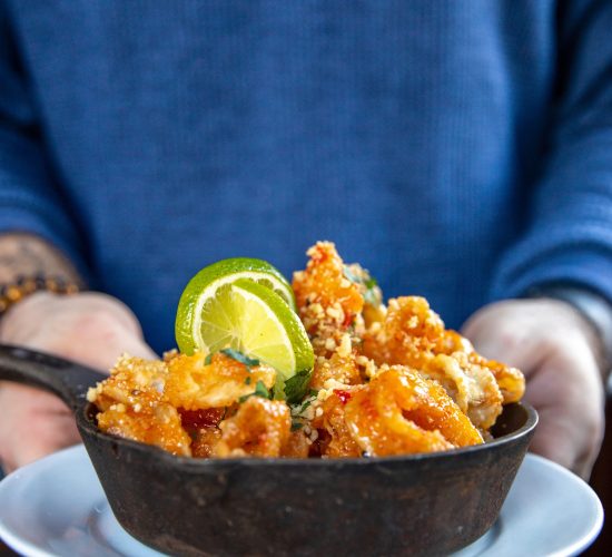 A person in a blue sweater holds a white plate with a small cast iron skillet filled with crispy fried shrimp, garnished with lime slices and herbs.