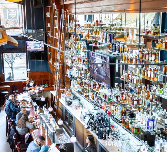 A bustling bar with patrons seated along a wooden counter. Shelves filled with a variety of liquor bottles cover the mirrored wall, extending to the ceiling. A bartender serves drinks, and a ladder provides access to the high shelves.