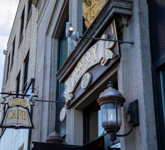 A city street features a building with two prominent signs: one reads "Library Cafe" with a hanging sign above the entrance, and the other is a decorative sign above the door. The entrance is framed by ornate stonework and exterior lights.