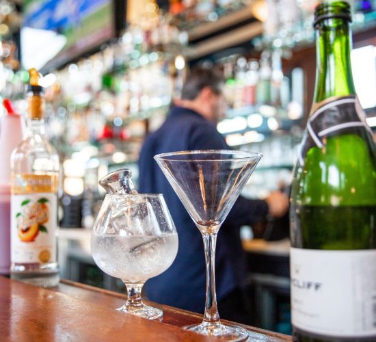 A bar scene with a variety of bottles and two glasses on a wooden counter: one is a stemmed glass with ice, and the other is a martini glass. A bartender works in the blurred background. Shelves of bottles are visible above.