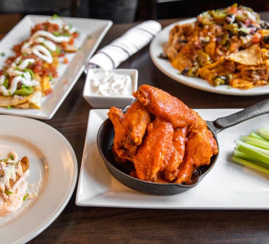 A variety of dishes on a table, including a skillet of buffalo wings with celery sticks, nachos topped with toppings and sour cream, and a salmon dish garnished with herbs and sauce. A small bowl of sour cream is also visible.