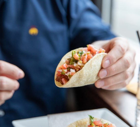 A person in a blue shirt is holding a taco filled with diced tomatoes and other toppings, with a plate of similar tacos on the table in front. The setting appears to be a casual dining restaurant.