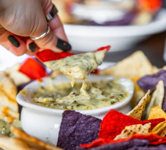 A hand dipping a tortilla chip into a bowl of creamy spinach artichoke dip. The bowl is surrounded by an assortment of colorful tortilla chips in red, yellow, and blue on a white plate.