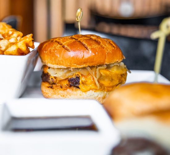 A close-up of a cheeseburger with a golden bun and melted cheese, garnished with a toothpick. Crispy waffle fries are visible in a white dish beside it. The background is softly blurred, highlighting the food.