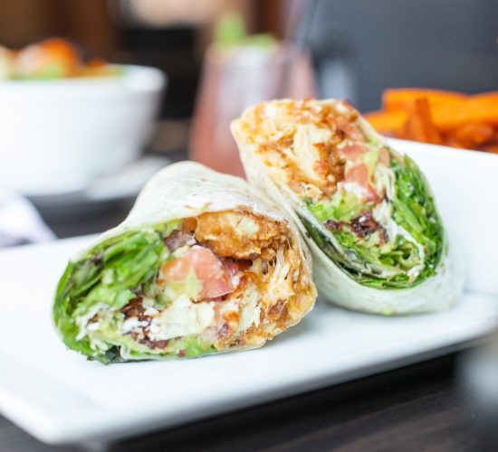 A close-up of a wrap sandwich cut in half, showcasing filling ingredients like crispy chicken, lettuce, tomatoes, and sauce on a white plate. In the background, there's a blurred image of a drink and a bowl of food.