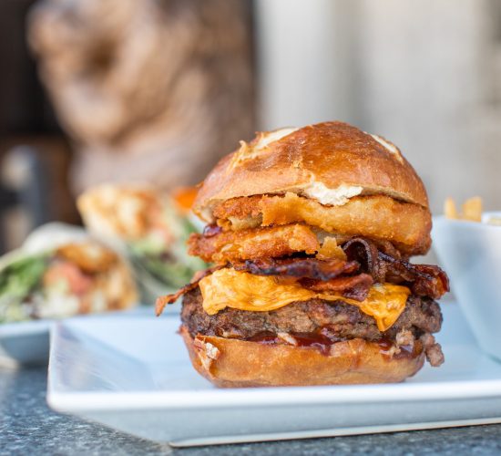 A close-up of a gourmet burger on a white plate, featuring a beef patty, melted cheese, crispy bacon, and an onion ring, topped with a brioche bun. In the background, there's a wrap on another plate and a dish of fries.