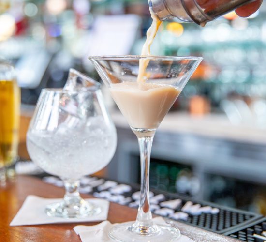 A creamy cocktail being poured from a shaker into a martini glass on a bar. Next to it, an empty snifter glass on a napkin. The bar's background is blurred, showing various bottles.