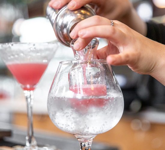 A bartender's hands are preparing a cocktail by pouring liquid from a shaker into a large glass with ice. A finished pink cocktail sits in a martini glass nearby, both on a bar counter.