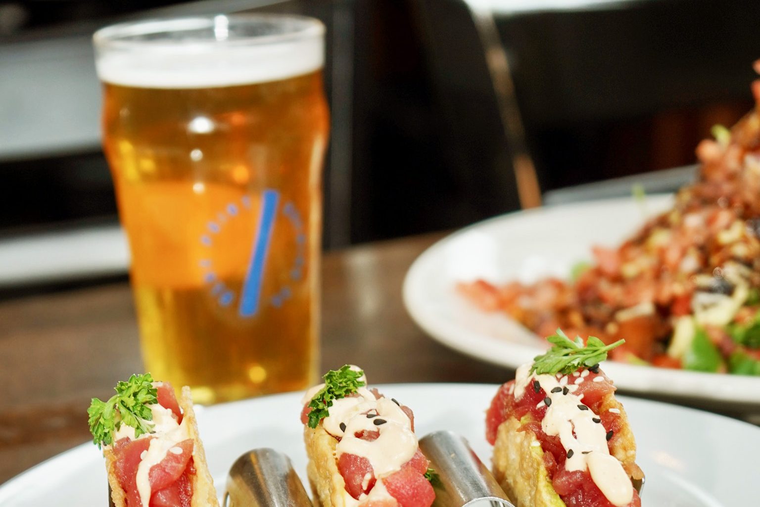 A glass of beer sits on a table next to a plate of mini tacos, filled with diced tuna, drizzled with creamy sauce, and garnished with herbs and sesame seeds. Menus featuring similar delightfully curated dishes are visible in the background.