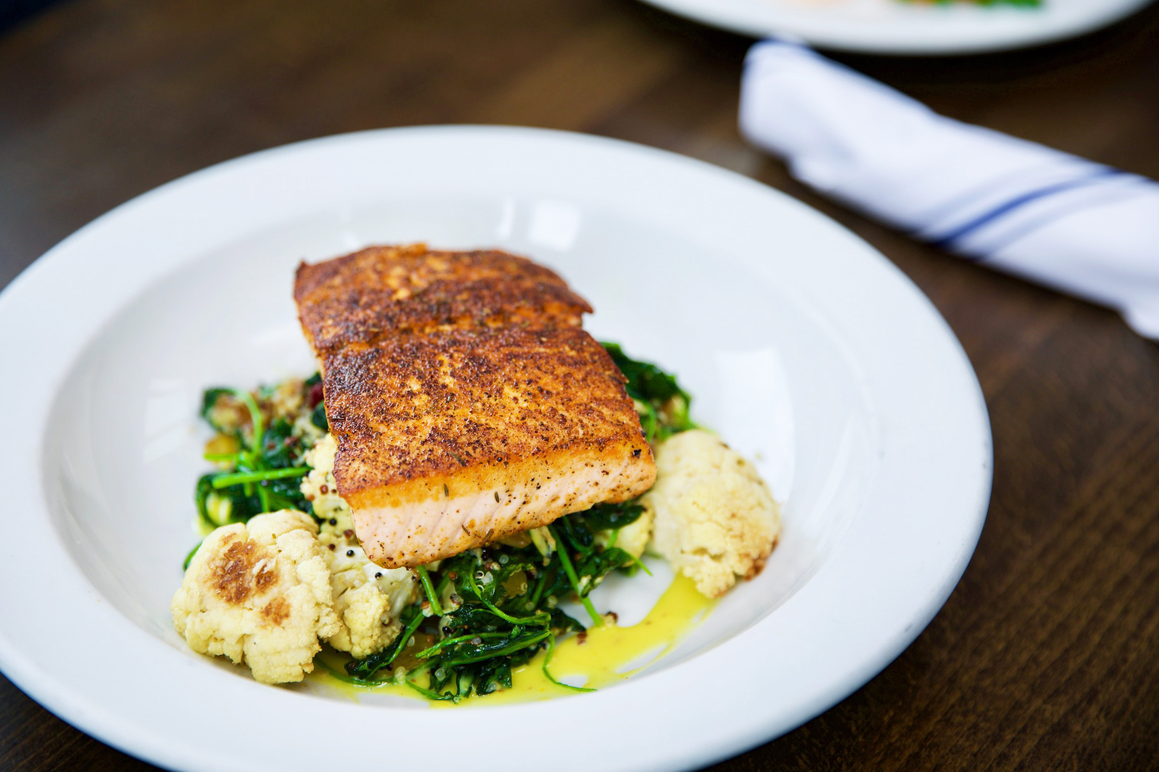 A plate featuring a seasoned, grilled salmon fillet atop a bed of sautéed greens and roasted cauliflower, drizzled with a light sauce, perfect for intimate events. A folded napkin is visible in the background, set on a wooden table.