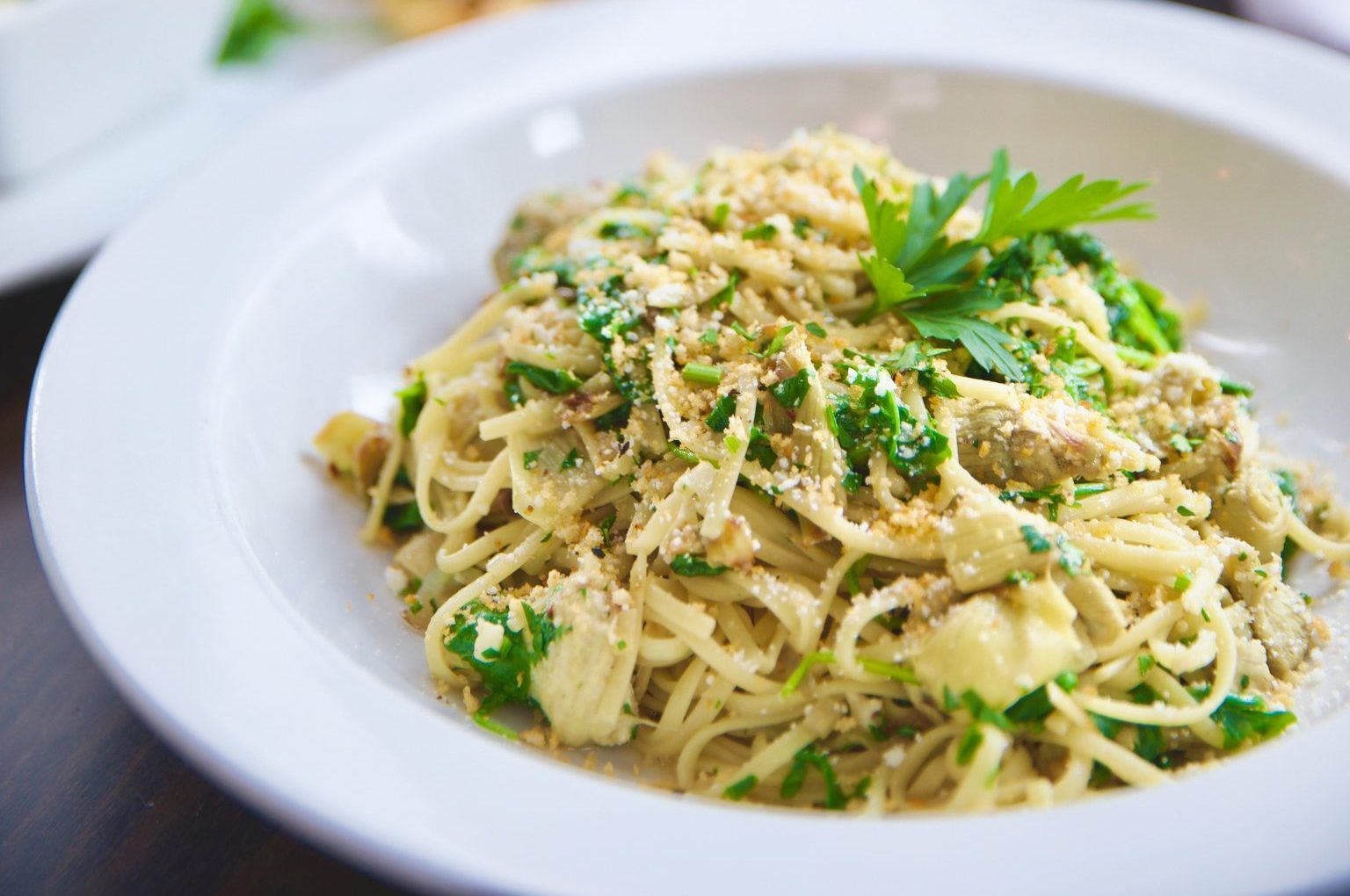 A close-up of a plate of spaghetti aglio e olio garnished with parsley, perfect for a luncheon. The pasta is mixed with garlic, olive oil, herbs, and sprinkled with Parmesan cheese. This delightful dish is served in a white bowl on a dark table surface.