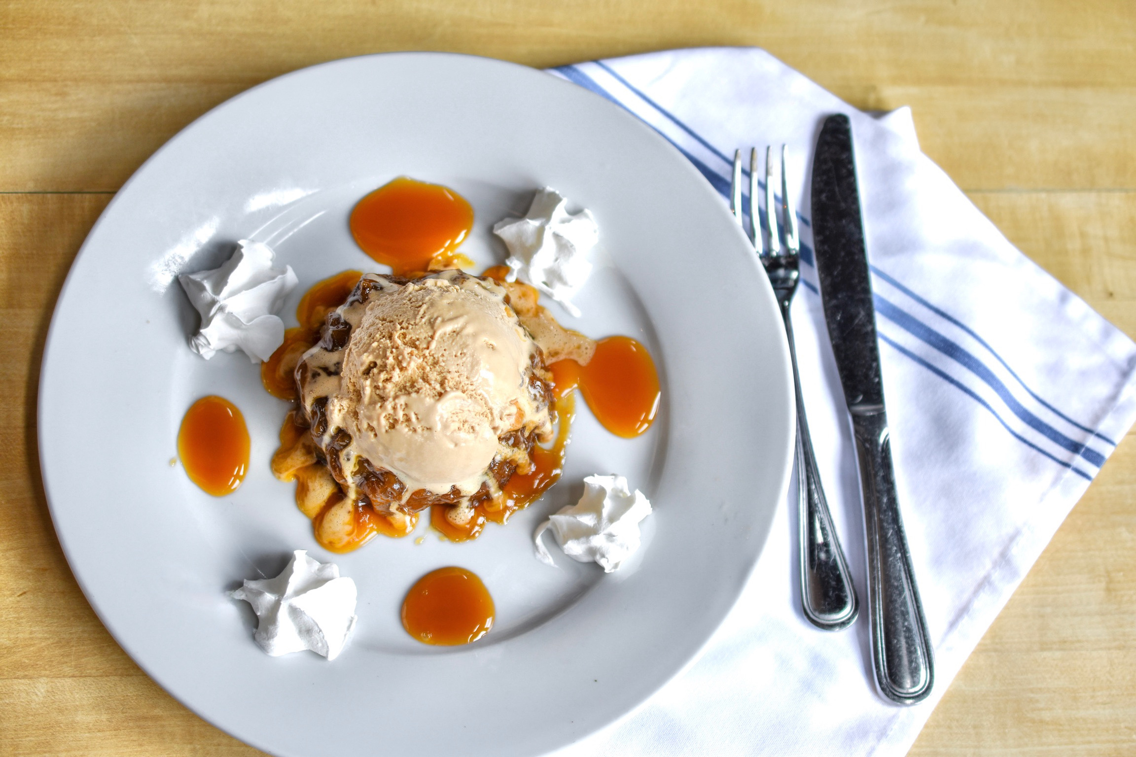 A dessert graces the menu with a scoop of ice cream drizzled in caramel sauce, set elegantly on a white plate. Dollops of whipped cream surround it, while a fork and knife rest on a napkin beside it on the rustic wooden table.