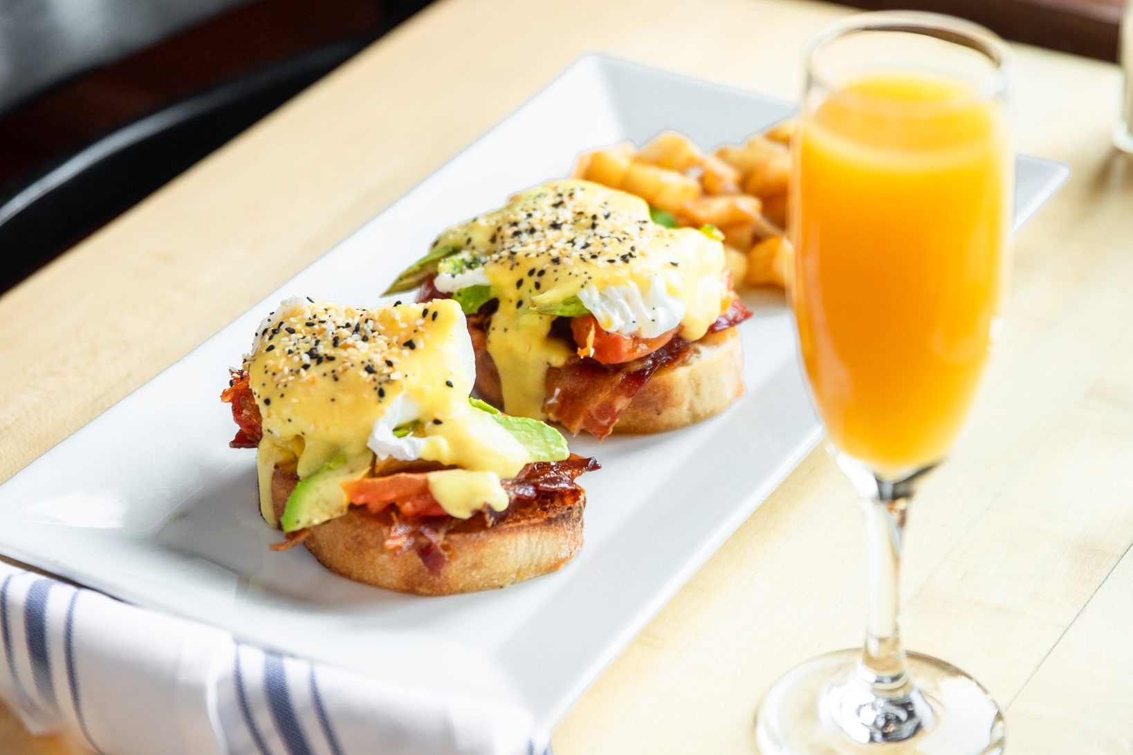 A plate of Eggs Benedict, perfect for any event, is topped with hollandaise sauce and garnished with seeds, served alongside diced potatoes. A glass of orange juice sits beside the plate on a wooden table.