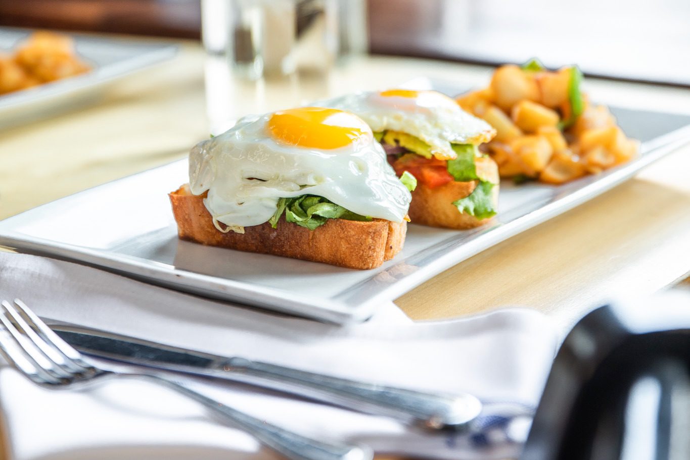 A plate from our menus features two slices of toasted bread topped with lettuce, tomato, and sunny-side-up eggs. On the side, enjoy diced potatoes garnished with herbs. The setting includes a fork and knife on a napkin.