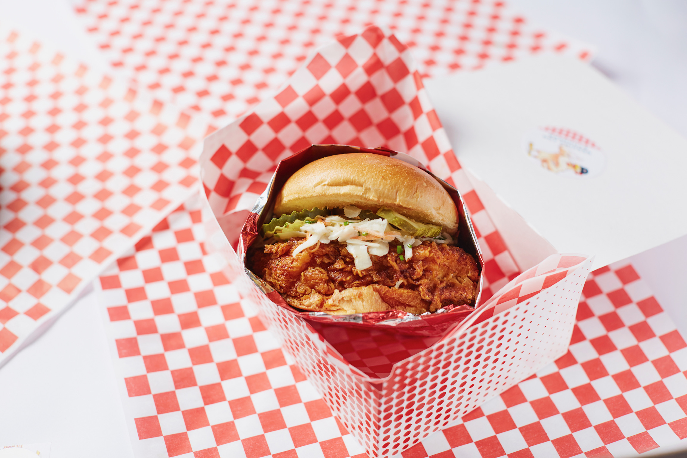 A crispy fried chicken sandwich with coleslaw and pickles is presented in red and white checkered paper, reminiscent of classic picnic menus, sitting in an open takeout box. The background features matching checkered paper.